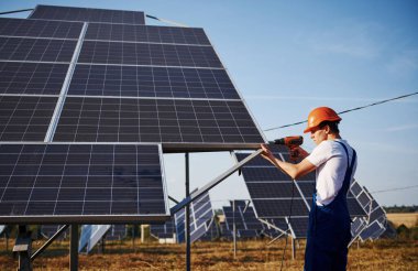 Kablosuz tornavida kullanıyor. Açık havada mavi üniformalı, güneşli bir günde güneş pilli bir erkek işçi..