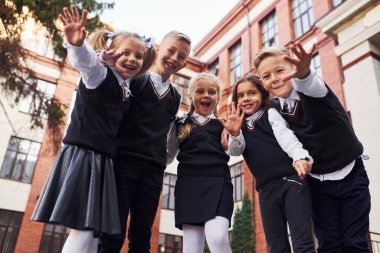 Eğleniyoruz ve birbirimizi kucaklıyoruz. Okul üniformalı bir grup çocuk eğitim binasının yanında açık havada beraberler..