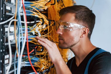 Koruyucu gözlüklü genç adam sunucu odasında internet cihazları ve kablolarla çalışıyor..