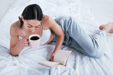 Stüdyoda elinde kitap ve bir bardak içkiyle uzanmış spor elbiseli pozitif gülümseyen bir kadın..