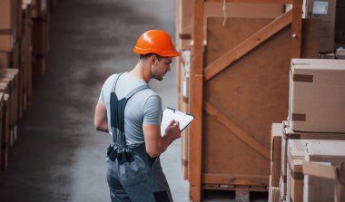 Üniformalı bir depo işçisi ve eldeki not defteri üretimi kontrol ediyor..