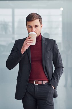 Lüks takım elbiseli ve resmi kıyafetli genç bir iş adamı. Bir bardak içkiyle ofisin içinde..