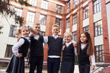 Eğleniyoruz ve birbirimizi kucaklıyoruz. Okul üniformalı bir grup çocuk eğitim binasının yanında açık havada beraberler..
