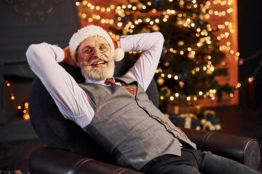 Süslü püslü, gri saçlı ve sakallı bir son sınıf öğrencisinin portresi süslü bir odada ve Noel şapkasıyla oturuyor..