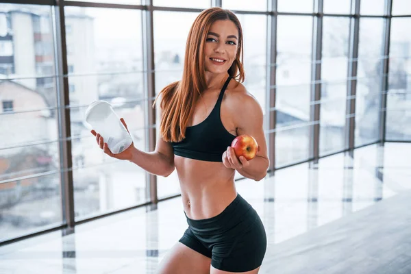 素敵な体を持つスポーティな女性は手の中に水と新鮮なリンゴのボトルと屋内スタンド — ストック写真