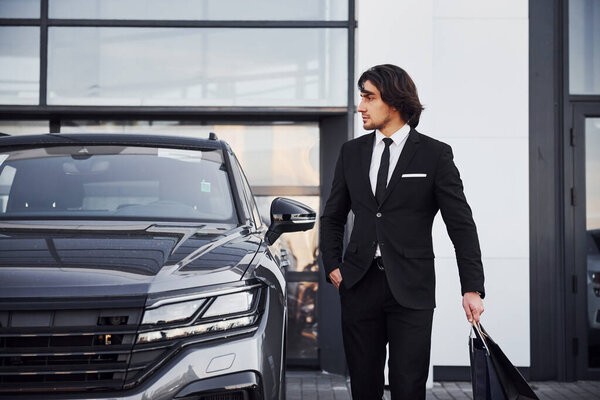 Portrait of handsome young businessman in black suit and tie outdoors near modern car and with shopping bags.