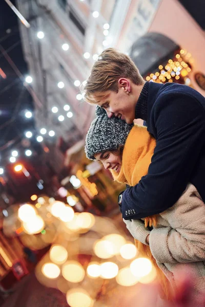 积极的年轻夫妇穿着温暖的衣服 在装饰过的圣诞大街上拥抱在一起 — 图库照片
