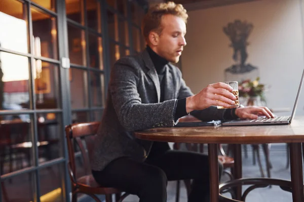 エレガントな若いです男でフォーマルな服でカフェに座っています彼のラップトップとアルコールのガラス — ストック写真