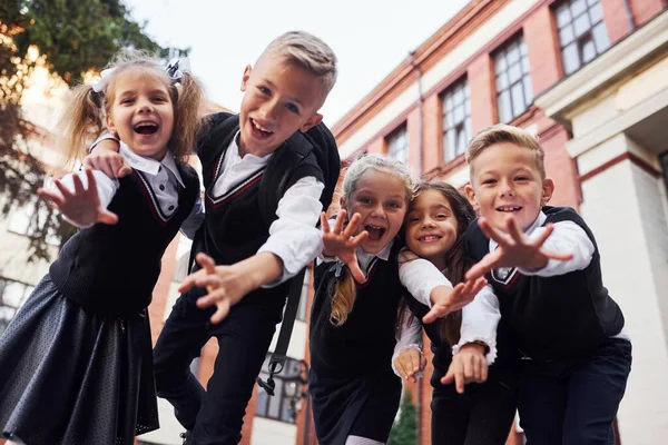 Eğleniyoruz ve birbirimizi kucaklıyoruz. Okul üniformalı bir grup çocuk eğitim binasının yanında açık havada beraberler..