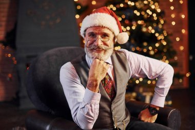 Süslü püslü, gri saçlı ve sakallı bir son sınıf öğrencisinin portresi süslü bir odada ve Noel şapkasıyla oturuyor..