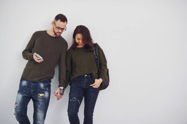 Sırt çantalı ve telefonlu neşeli çoklu etnik çift stüdyoda beyaz arka plana karşı yan yana duruyorlar..