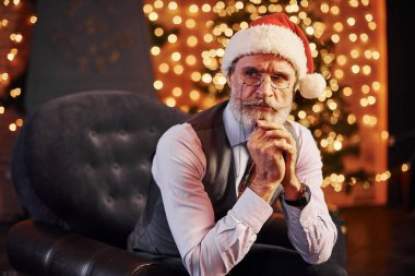 Süslü püslü, gri saçlı ve sakallı bir son sınıf öğrencisinin portresi süslü bir odada ve Noel şapkasıyla oturuyor..