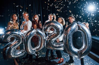 Konfeti havada. 2020 yılını kutlamak için ellerinde balonlar ve içkilerle neşeli bir grup insan.