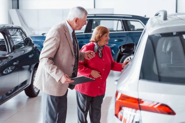 Resmi kıyafetli yaşlı bir adam ve otomobil seçerken yanında not defteri olan bir kadın..