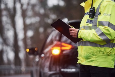 Yeşil üniformalı erkek polis memuru arabanın yanında not defteriyle dikiliyor..