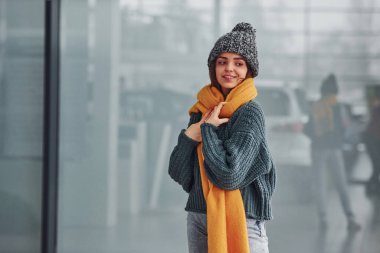 Sarı eşarplı, kalın elbiseli, güzel, neşeli bir kız. Arka planda yansımaları olan kapalı bir yerde duruyor..