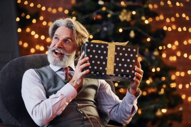 Gri saçlı ve sakallı son sınıf öğrencisinin portresi hediye kutusuyla süslenmiş Noel odasında..