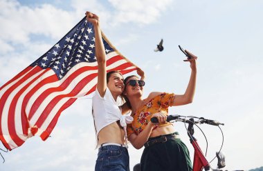Bulutlu gökyüzüne karşı. İki vatansever kadın ellerinde bisiklet ve Amerikan bayrağı ile selfie çekiyorlar..