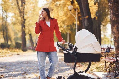 Kırmızı ceketli anne, çocuğuyla birlikte sonbahar zamanı parkta yürüyüş yapar ve sigara içer..