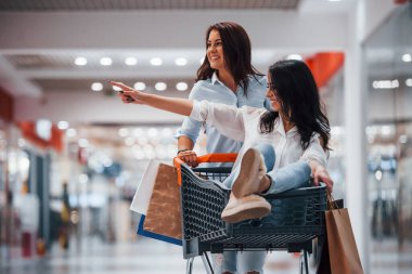 İki genç kadın süpermarkette alışveriş sepetinde koşup eğleniyorlar..