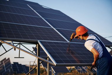 Kablosuz tornavida kullanıyor. Açık havada mavi üniformalı, güneşli bir günde güneş pilli bir erkek işçi..