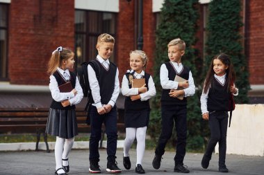 Okul üniformalı bir grup çocuk eğitim binasının yanında açık havada beraberler..