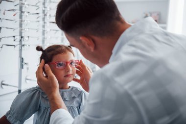 Beyaz önlüklü genç bir çocuk doktoru küçük kız için yeni gözlük almasına yardım ediyor..