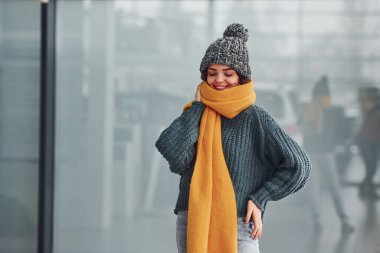 Sarı eşarplı, kalın elbiseli, güzel, neşeli bir kız. Arka planda yansımaları olan kapalı bir yerde duruyor..