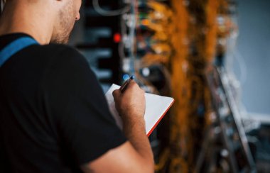 Elinde not defteri olan üniformalı genç adam sunucu odasında internet ekipmanı ve kablolarla çalışıyor..