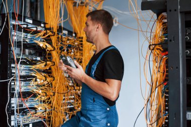 Ölçüm cihazlı üniformalı genç adam sunucu odasında internet ekipmanı ve kablolarla çalışıyor..