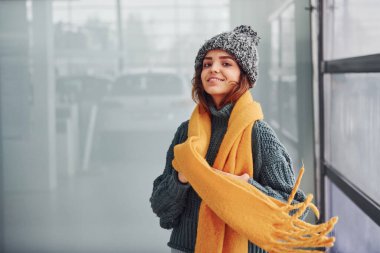 Sarı eşarplı, kalın elbiseli, güzel, neşeli bir kız. Arka planda yansımaları olan kapalı bir yerde duruyor..