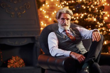 Gri saçlı ve sakallı son sınıf öğrencisinin portresi süslü Noel odasında sandalyede oturuyor..