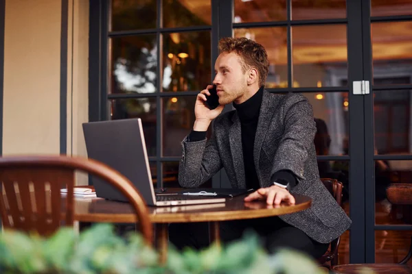 エレガントな若いです男でフォーマル服で座ってカフェで彼のラップトップと電話で話す — ストック写真