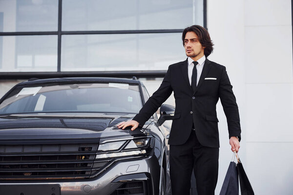 Portrait of handsome young businessman in black suit and tie outdoors near modern car and with shopping bags.