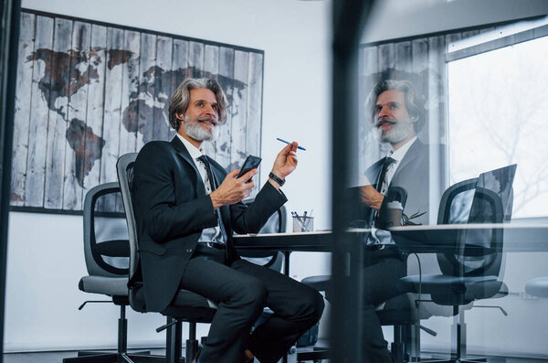 В офисе пожилой бизнесмен с седыми волосами и бородой в формальной одежде.