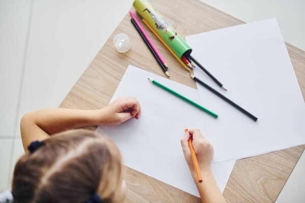 Вид сверху маленькой девочки с ручкой и пустым листом бумаги на столе.