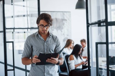 Elinde kalem, elinde not defteri olan gözlüklü bir adam karşıdaki ofiste duruyor..