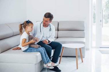 Beyaz üniformalı bir erkek doktor klinikte küçük bir kızla oturuyor..