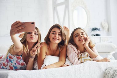 Pembe telefon kullanarak selfie çekerek. Mutlu bayan arkadaşlar yatak odasında pijama partisinde iyi vakit geçiriyorlar..