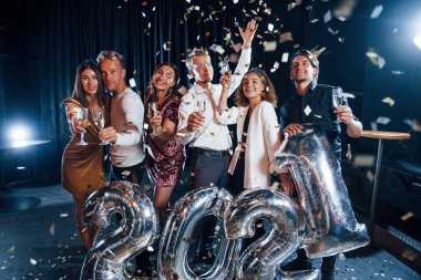 Konfeti havada uçuşuyor. Yeni yıl kutlamaları için ellerinde balonlar ve içkilerle neşeli bir grup insan..