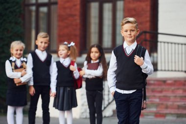 Okul üniformalı bir grup çocuk eğitim binasının yanında kameraya poz veriyor..