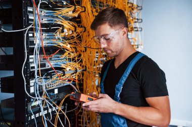 Ölçüm cihazlı üniformalı genç adam sunucu odasında internet ekipmanı ve kablolarla çalışıyor..