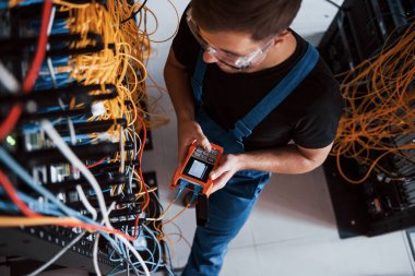 Sunucu odasında internet ekipmanı ve kablolarla çalışan ölçüm cihazıyla üniformalı genç adamın en iyi görüntüsü.