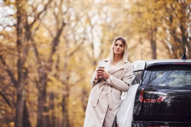 Ellerinde bir bardak içkiyle. Kızın arabayla sonbahar gezisi var. Ormanda yepyeni bir otomobil..