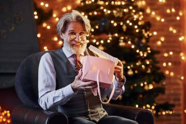 Gri saçlı ve sakallı son sınıf öğrencisinin portresi hediye kutusuyla süslenmiş Noel odasında..