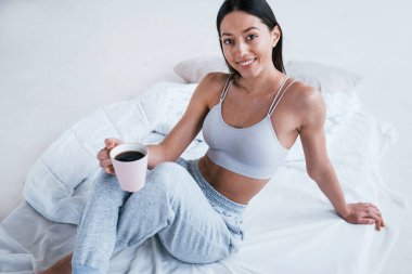 Sportif giysiler içindeki pozitif gülümseyen kadın bir bardak içkiyle stüdyoda oturuyor..