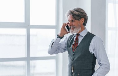 Gülümseyen, ceketli ve kravatlı mutlu bir adam. Kapalı bir odada, pencerenin önünde telefonla konuşuyor..