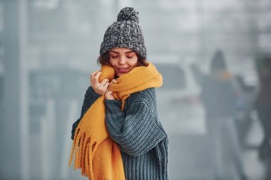 Sarı eşarplı, kalın elbiseli, güzel, neşeli bir kız. Arka planda yansımaları olan kapalı bir yerde duruyor..
