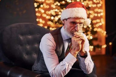 Süslü püslü, gri saçlı ve sakallı bir son sınıf öğrencisinin portresi süslü bir odada ve Noel şapkasıyla oturuyor..