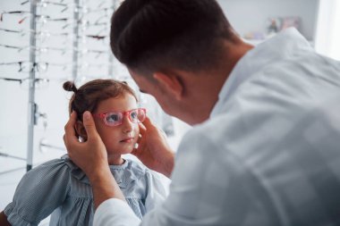 Beyaz önlüklü genç bir çocuk doktoru küçük kız için yeni gözlük almasına yardım ediyor..
